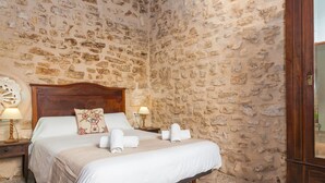 Un dormitorio con pared de piedra, estructura de cama de madera, ropa de cama blanca y dos lámparas de noche.