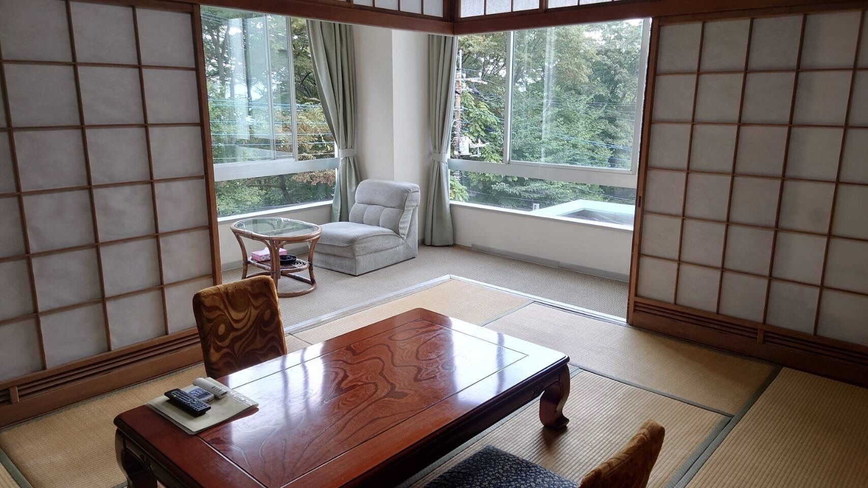 A traditional Japanese room with tatami flooring, a wooden table, a cushioned chair, and a view of greenery through large windows.