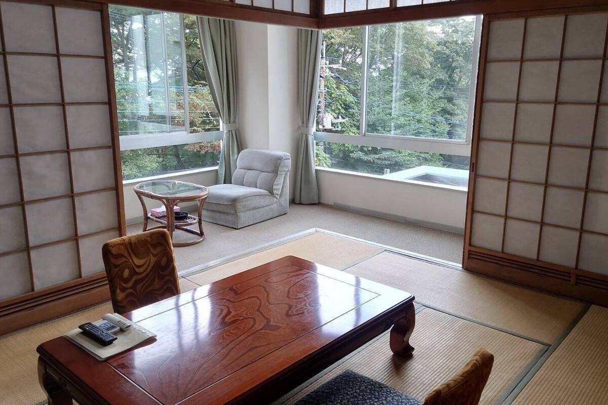 Um quarto japonês tradicional com tatames, uma mesa de madeira, uma poltrona acolchoada e vista para o verde através de grandes janelas.