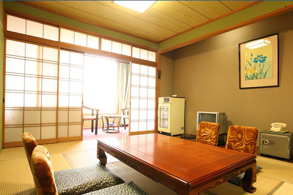 Um quarto japonês tradicional com tatames, uma mesa de madeira, cadeiras, uma televisão e um telefone.
