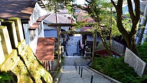 Una escalera de piedra conduce a un edificio tradicional japonés con techo de tejas, rodeado de vegetación y un pequeño santuario.