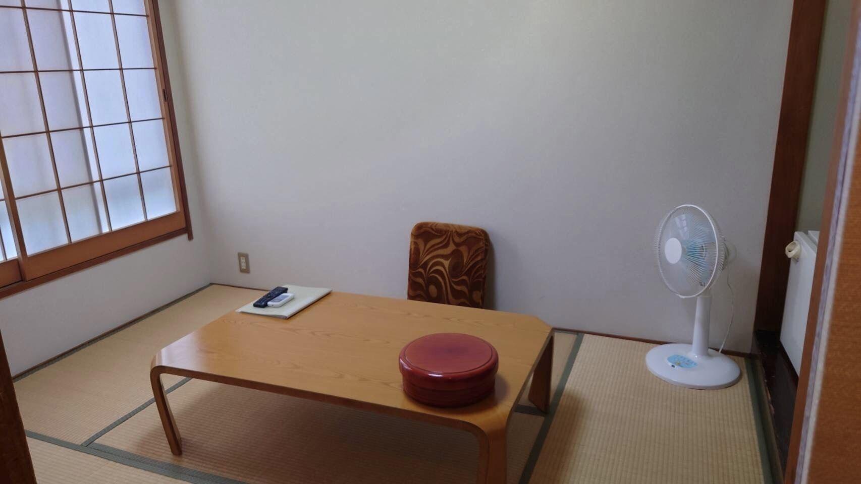 A room with tatami flooring, a wooden table with a red container, a chair, a fan, and a window with a grid pattern.