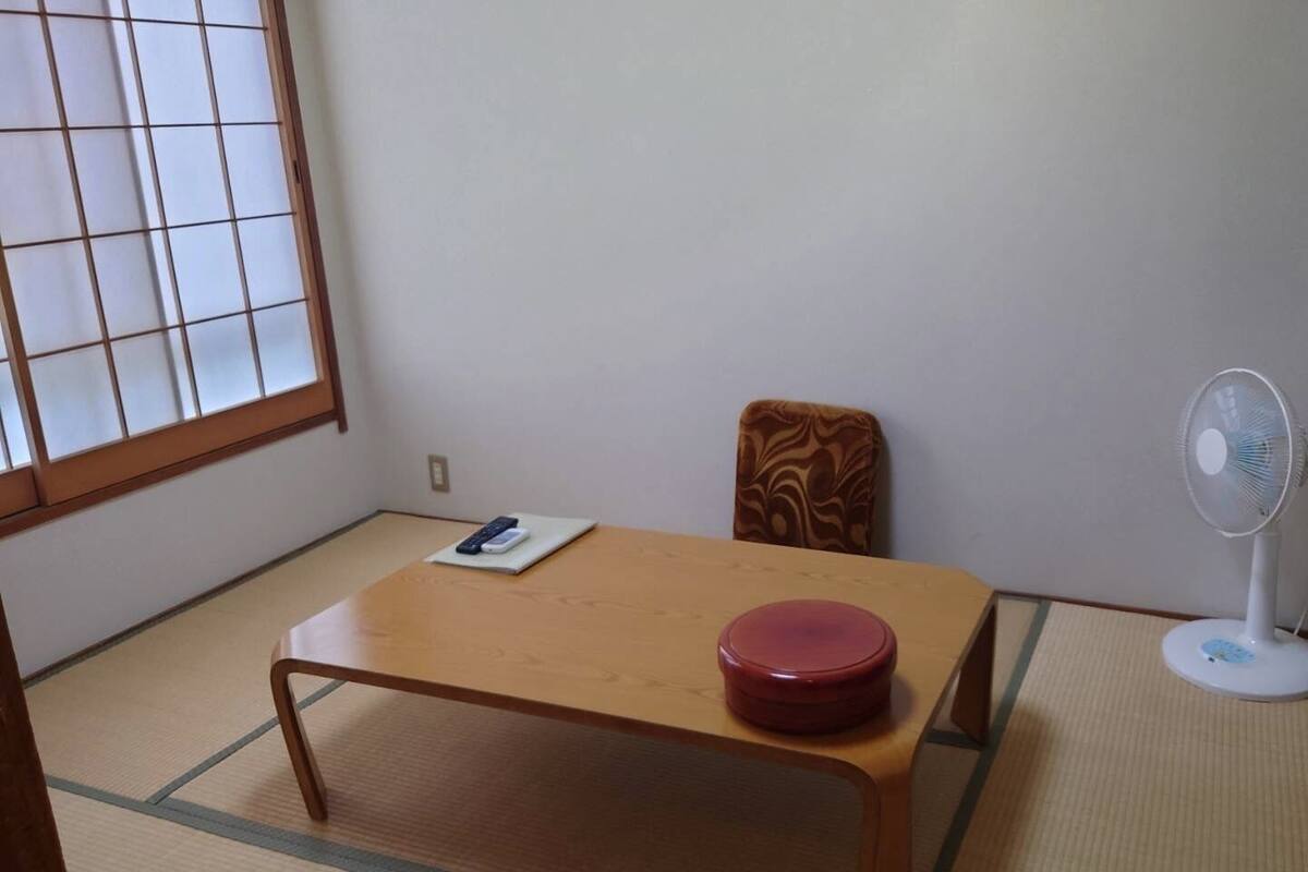 Quarto com piso de tatame, uma mesa de madeira com um recipiente vermelho, uma cadeira, um ventilador e uma janela com padrão quadriculado.