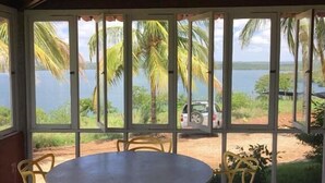 Ein überdachter Außenbereich mit einem runden Tisch und Stühlen, große Fenster mit Blick auf Palmen und ein Gewässer.