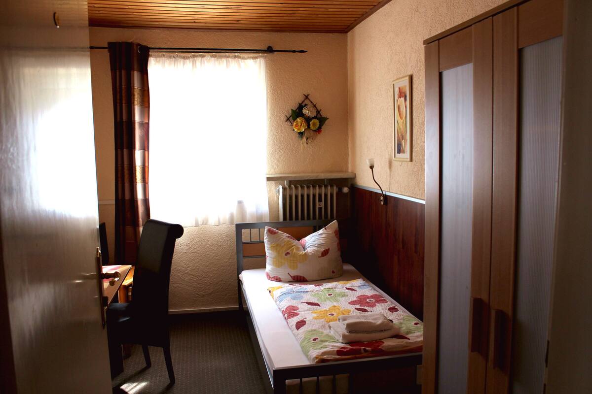 Ein kleines, ordentlich eingerichtetes Schlafzimmer mit einem Einzelbett, einem Sessel, einer Heizung und einem Fenster mit Vorhängen.
