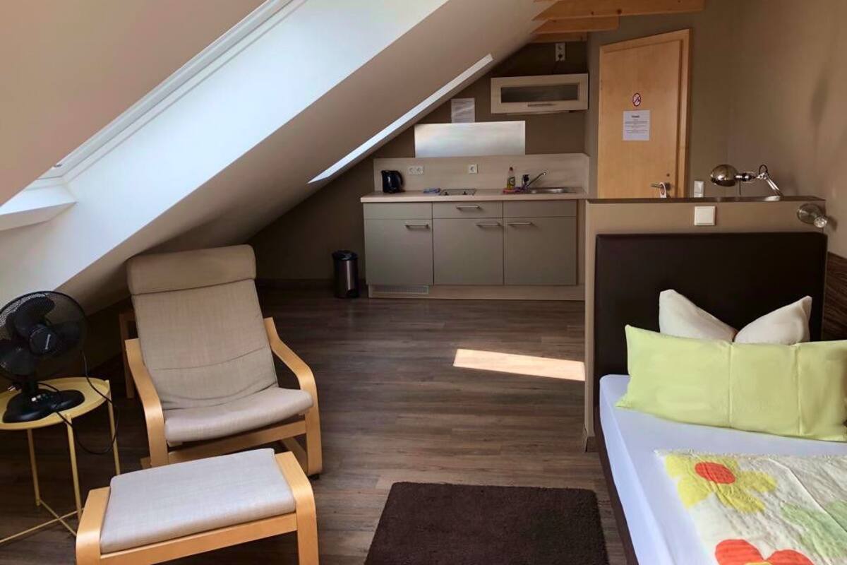 Ein kleines Zimmer mit einem Bett, einem Sessel, einem kleinen Tisch, einem Ventilator und einer Küchenzeile im Hintergrund.
