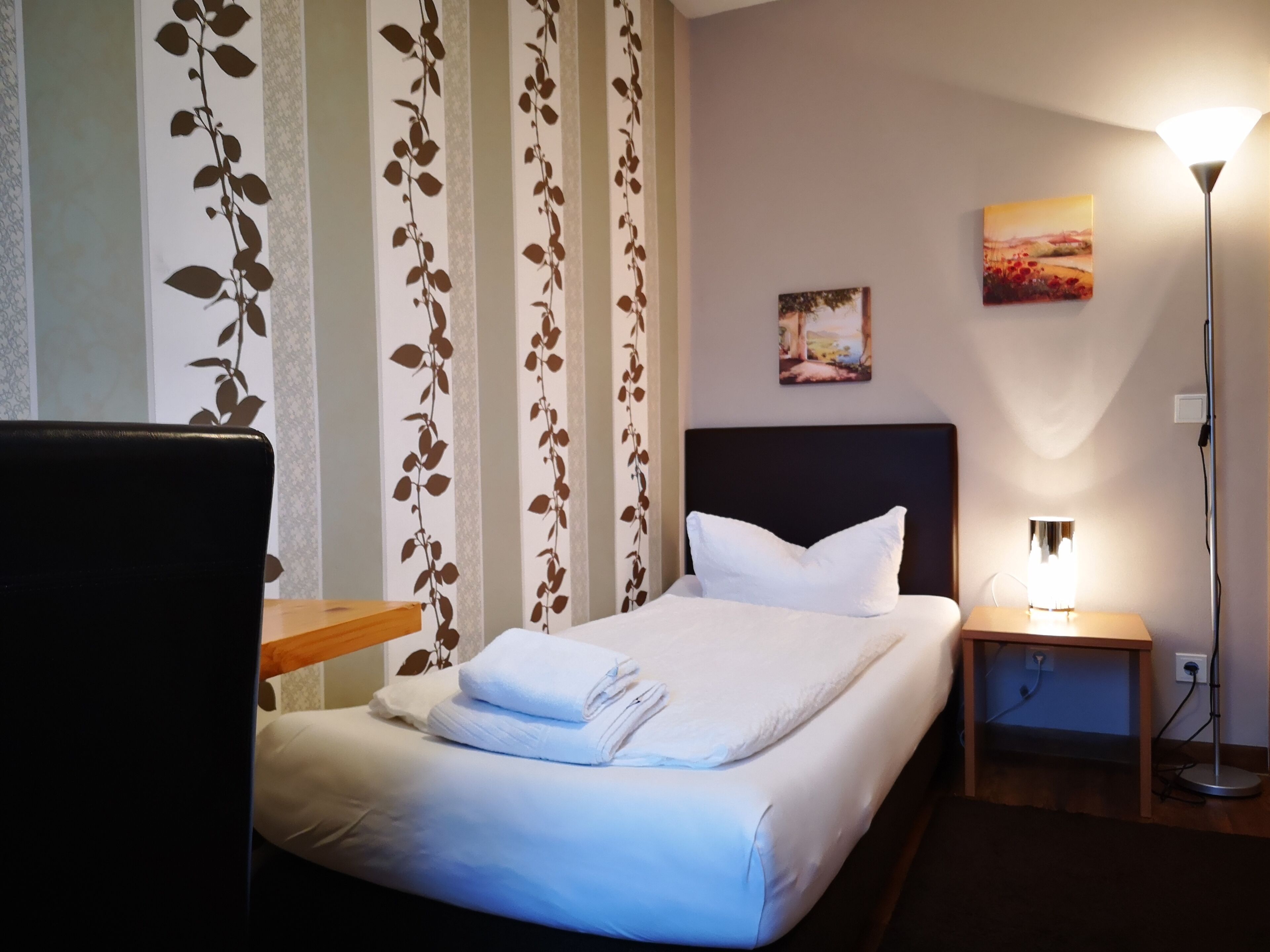 A neatly made bed with white linens and a pillow, a bedside table with a lamp, and a wall with a floral pattern and a painting.
