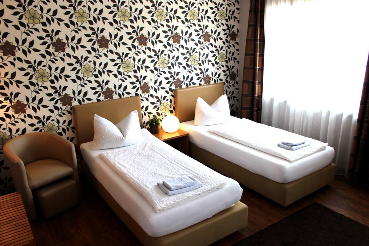Ein Hotelzimmer mit zwei Einzelbetten, einem Sessel, einem kleinen Tisch und floralem Wandpapier.
