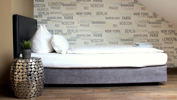 A bed with a grey mattress and white pillows, a round metallic side table with a plant, and a wall with city names in large letters. - greenpartment Boardinghouse Hotel Ingolstadt Friedrichshofen (Ingolstadt)