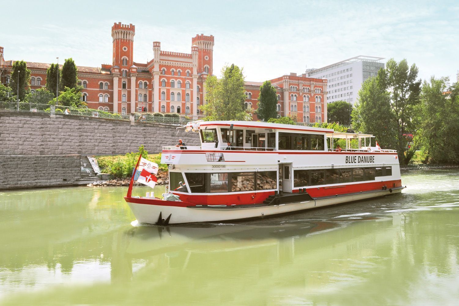 Ein Flussdampfer namens „BLUE DANUBE“ mit roter und weißer Außenbordwand, liegt an einem historischen Gebäude mit Türmen festgemacht.