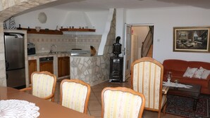 A kitchen with a stone backsplash, wooden cabinets, and a built-in stove. There is a dining area with a table and chairs, and a living room with a sofa and a painting on the wall. - Villa Mara (Slivno)