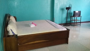 Una cama individual con estructura de madera, un colchón blanco y una toalla rosa. Hay una mesita con un ventilador y un televisor.