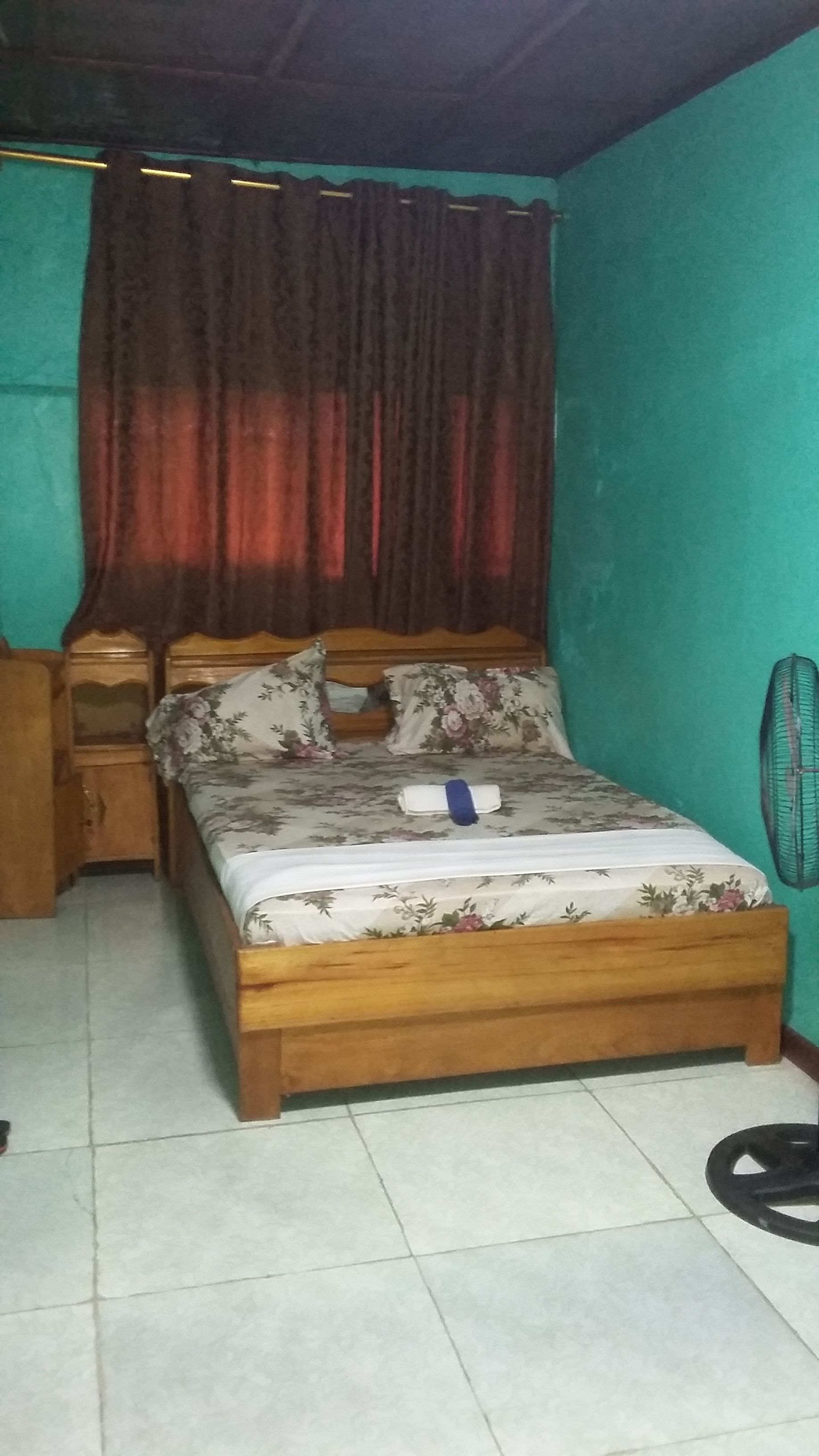 Een slaapkamer met een houten bed, bloemenbeddengoed, een nachtkastje, een bureau, een stoel en een ventilator.
