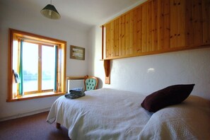 Quarto com cama, janela com cortinas, uma cadeira e teto de madeira.