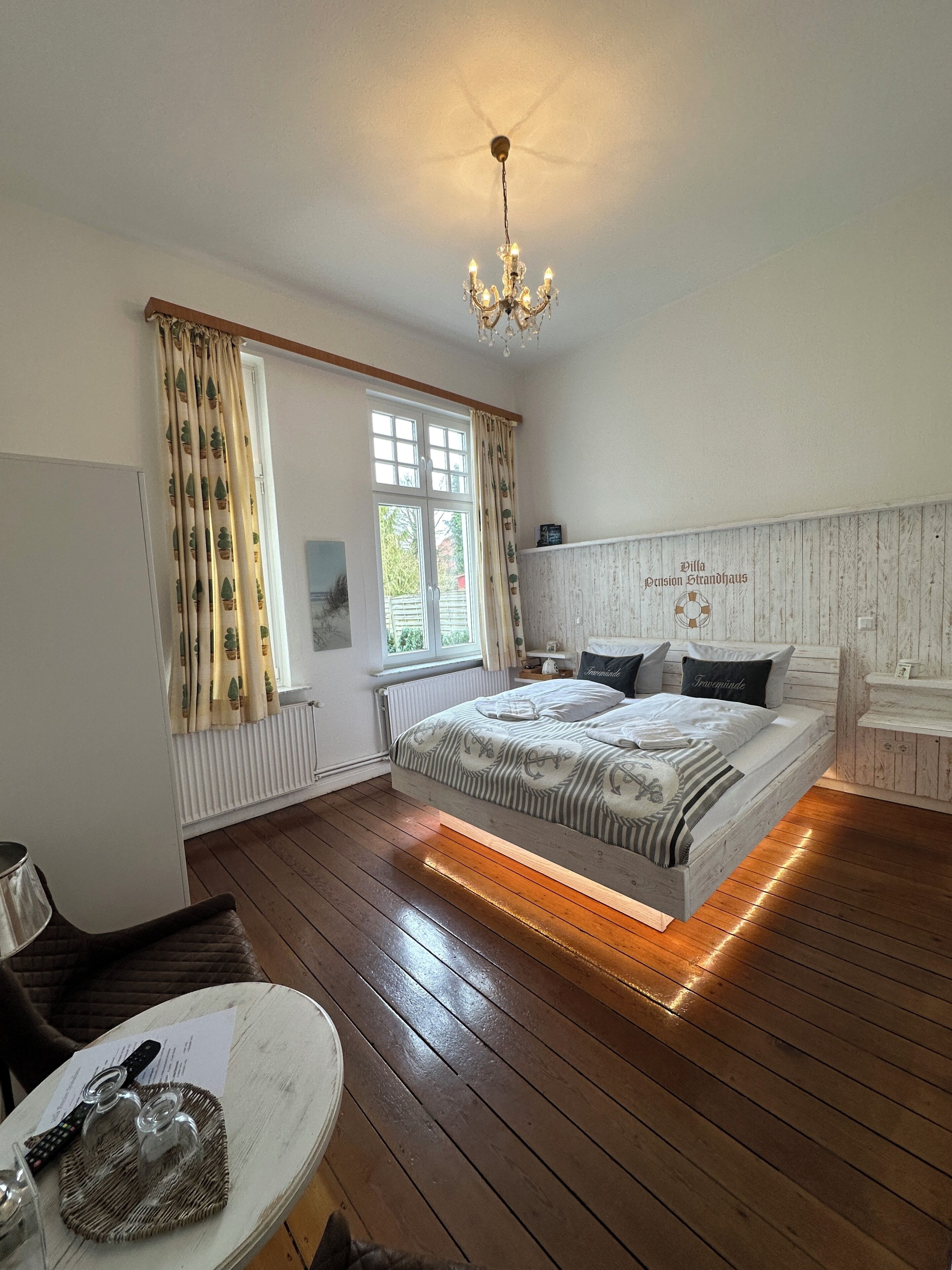 Une chambre avec un plancher en bois, un lit doté d’une tête de lit blanche, un lustre et une fenêtre avec des rideaux.