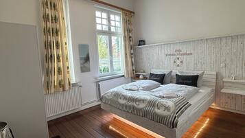 Une chambre avec un plancher en bois, un lit doté d’une tête de lit blanche, un lustre et une fenêtre avec des rideaux.