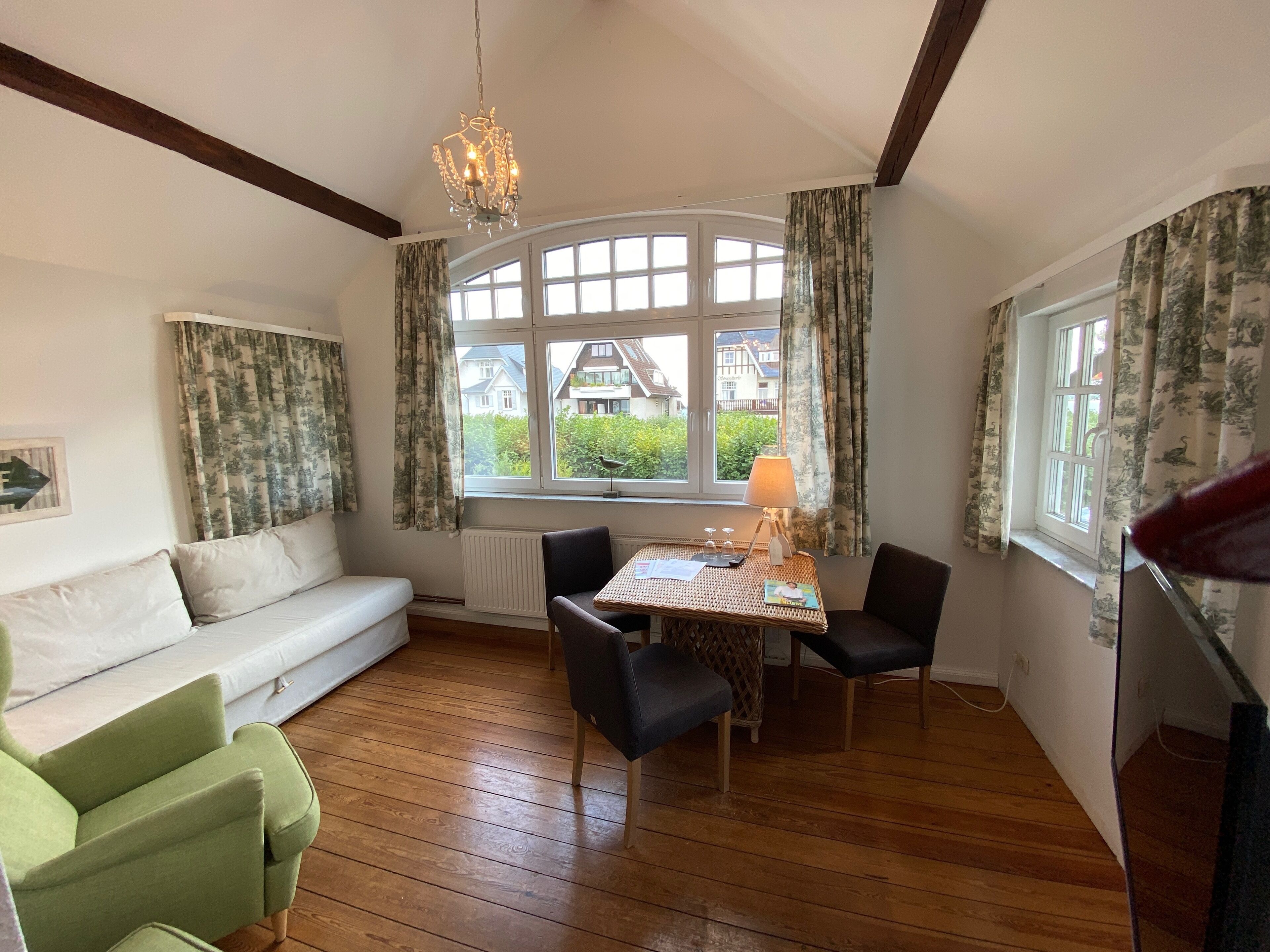 A cozy room with a wooden floor, a white sofa, a dining table with chairs, and large windows with curtains.