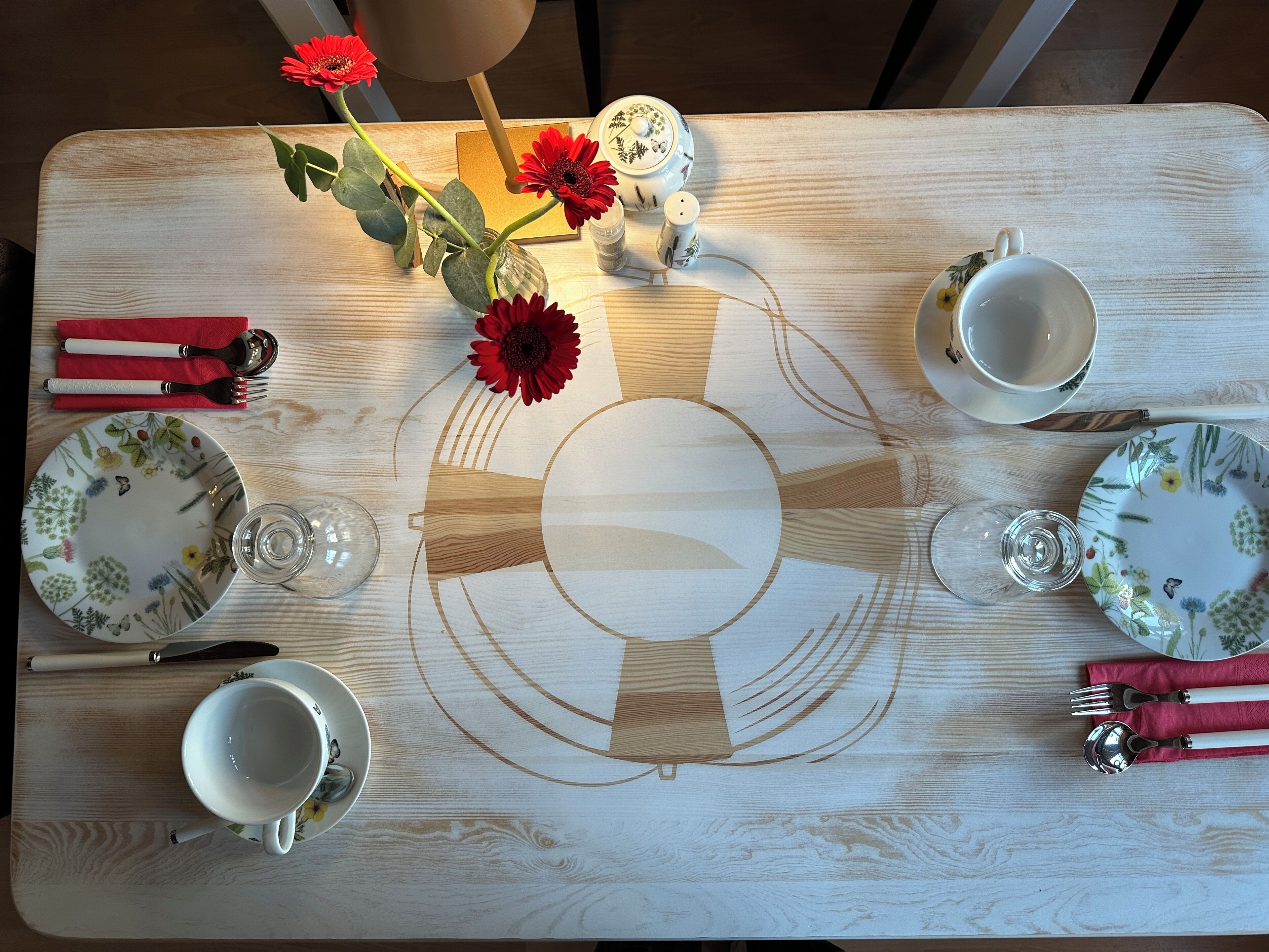A dining table with floral-patterned plates, cutlery, and a vase with red flowers.