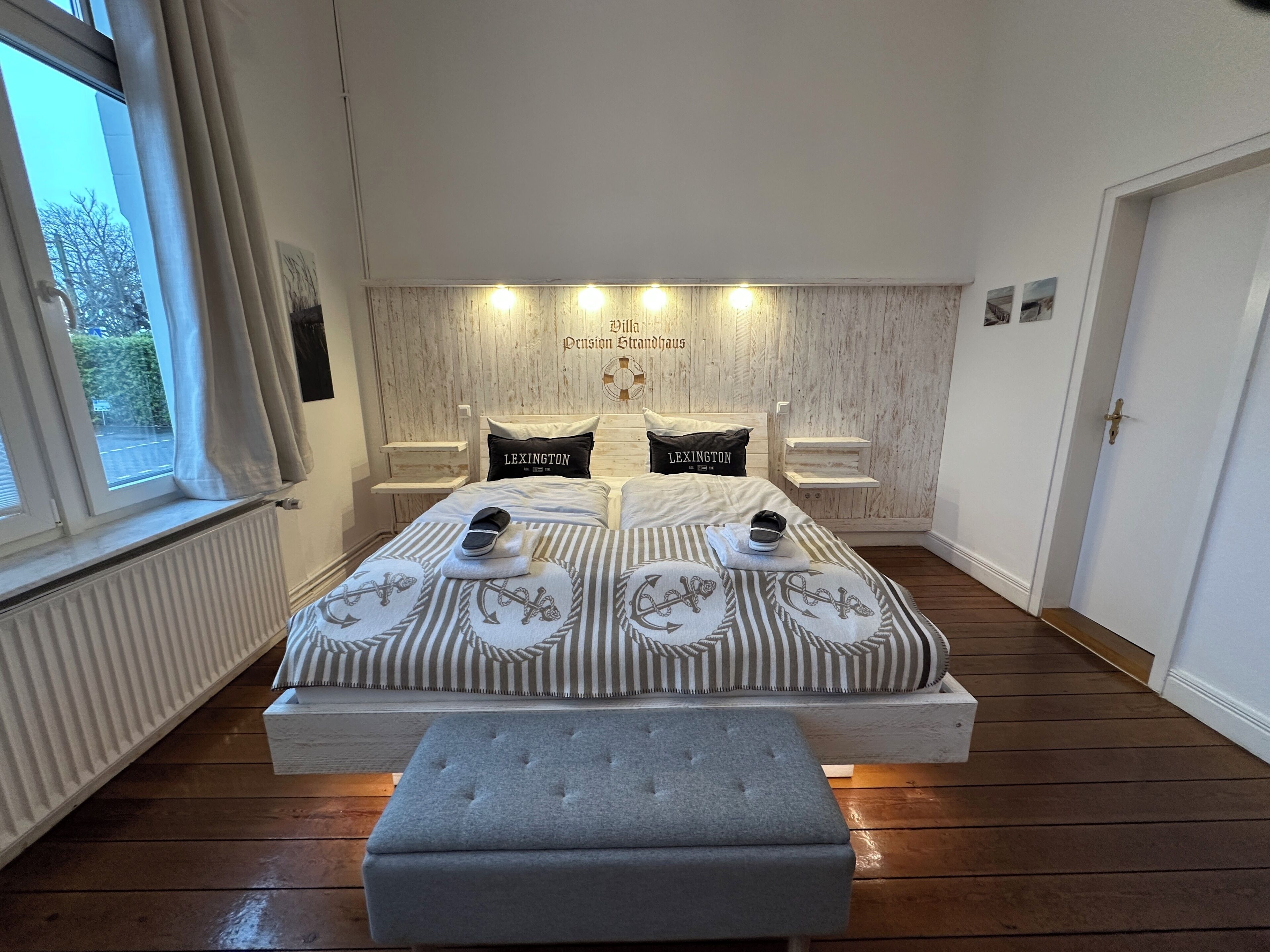A double bed with striped bedding, two pillows with 'LEXINGTON' text, a bedside table, a bench, and a view of the outside through a window.