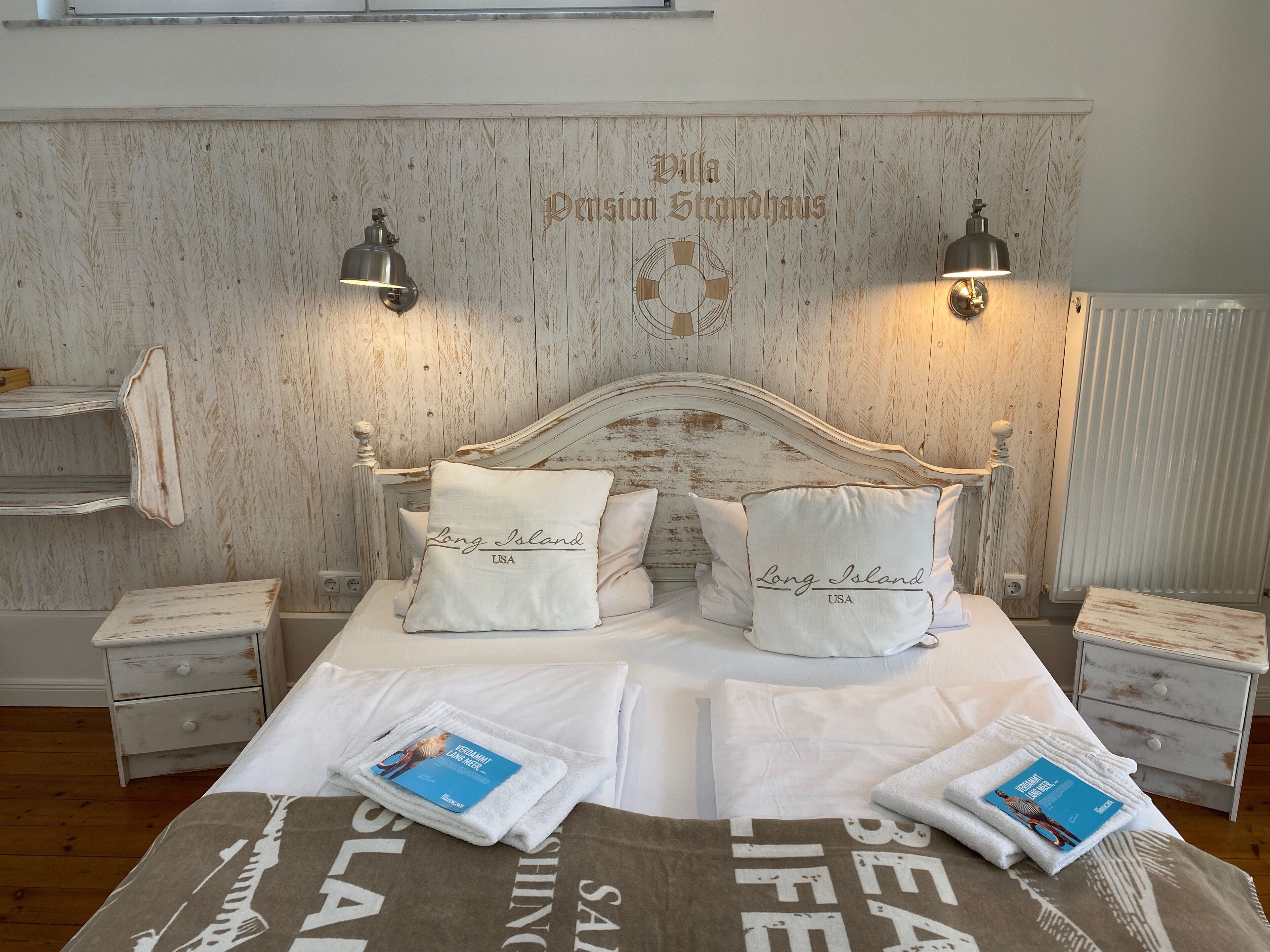 A bedroom with a wooden headboard, two bedside lamps, and two pillows with 'Long Island USA' text. There are two bedside tables with books and a towel.