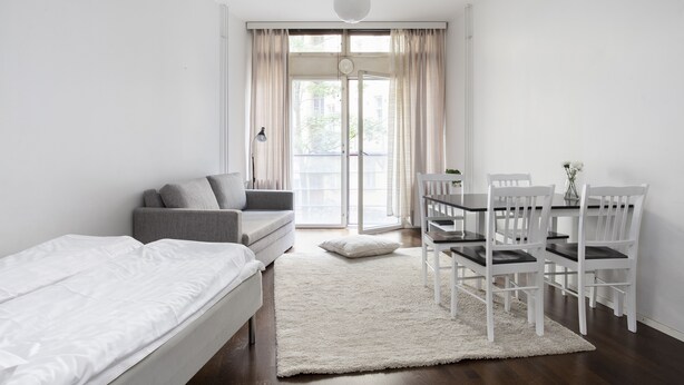 A modern, minimalist bedroom with a bed, a grey sofa, a dining table with white chairs, and a large window with curtains. - 2ndhomes Kluuvi Studios (Helsinki)