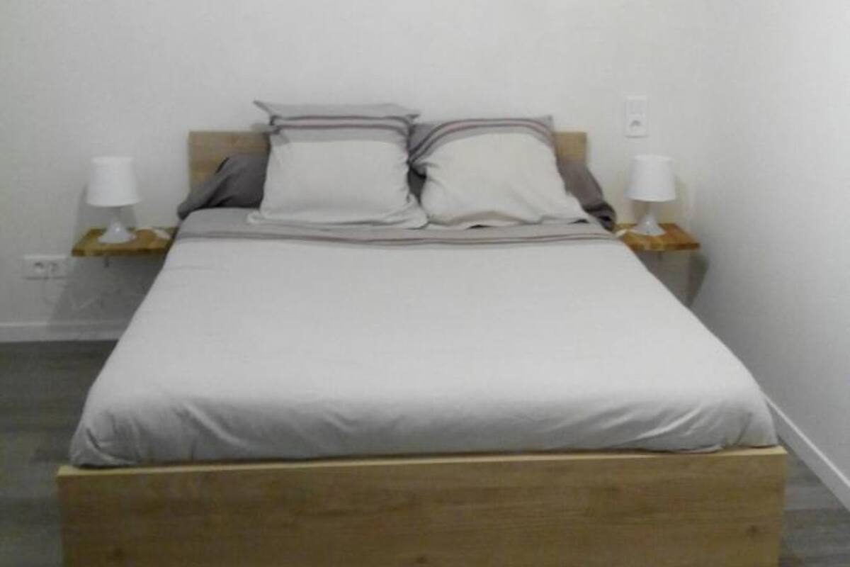 Une chambre avec un lit simple, une tête de lit en bois, du linge de lit blanc, deux tables de chevet avec des lampes et un mur blanc uni.