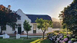 A white building with a dark roof, surrounded by a well-maintained lawn and a variety of flowering plants. - Cape Dutch at Keerweder (Franschhoek)