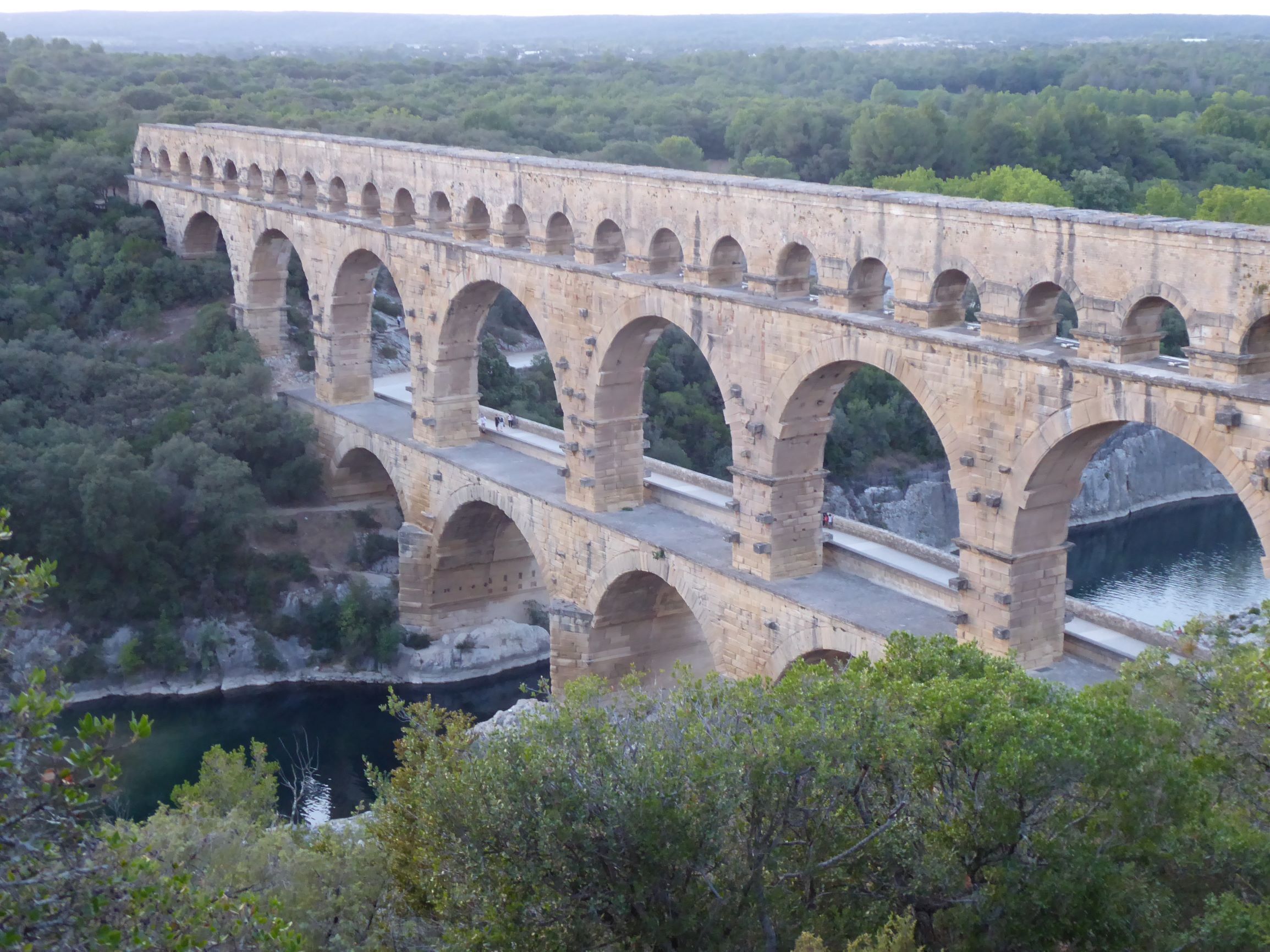 A stone aqueduct with multiple arches spanning a river, surrounded by dense greenery.