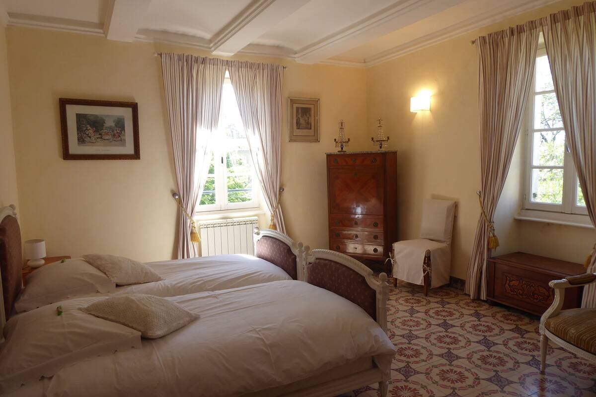 Quarto com duas camas, cômoda, cadeira e janela com cortinas.