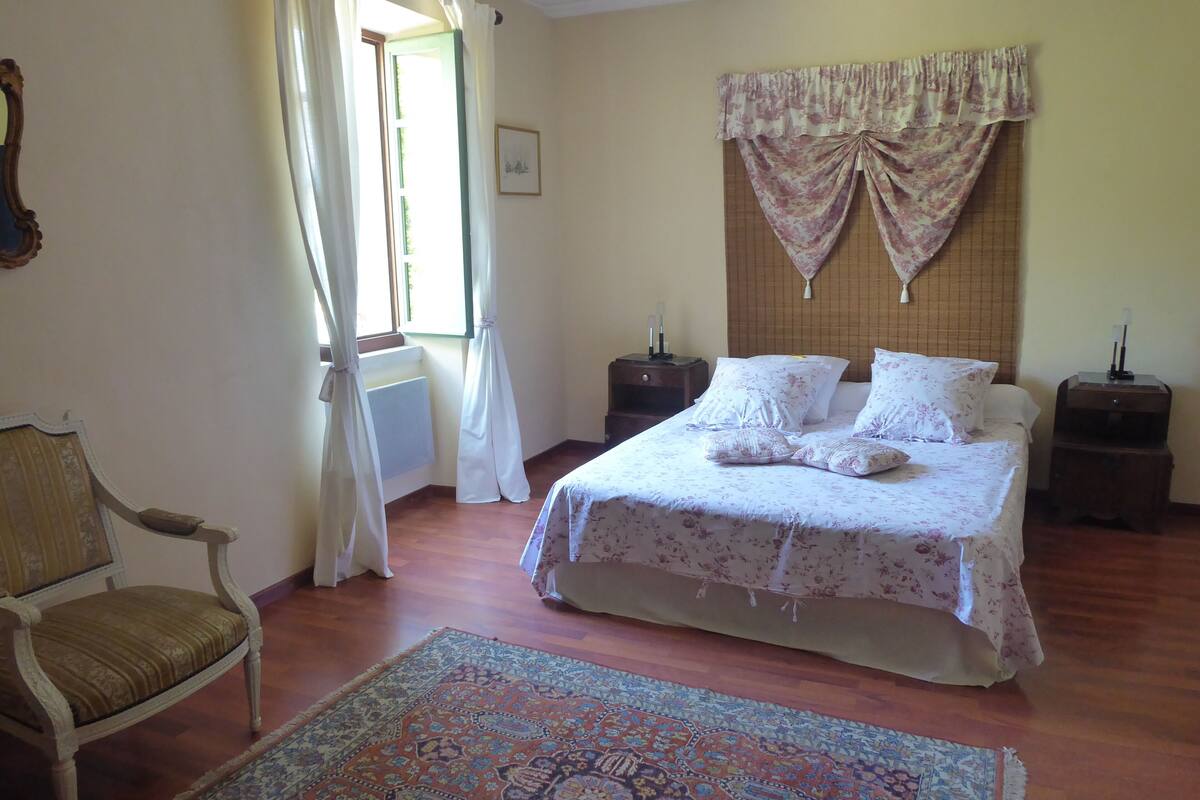 Quarto com cama, cadeira, mesa de cabeceira, cortinas numa janela e tapete estampado.