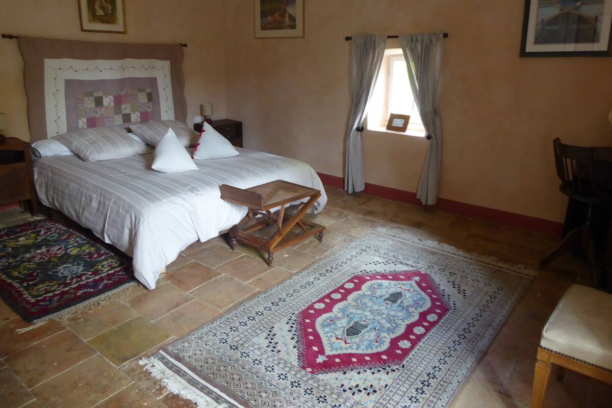 Quarto com cama, uma mesinha de madeira, cadeira, tapete estampado e janela com cortinas.