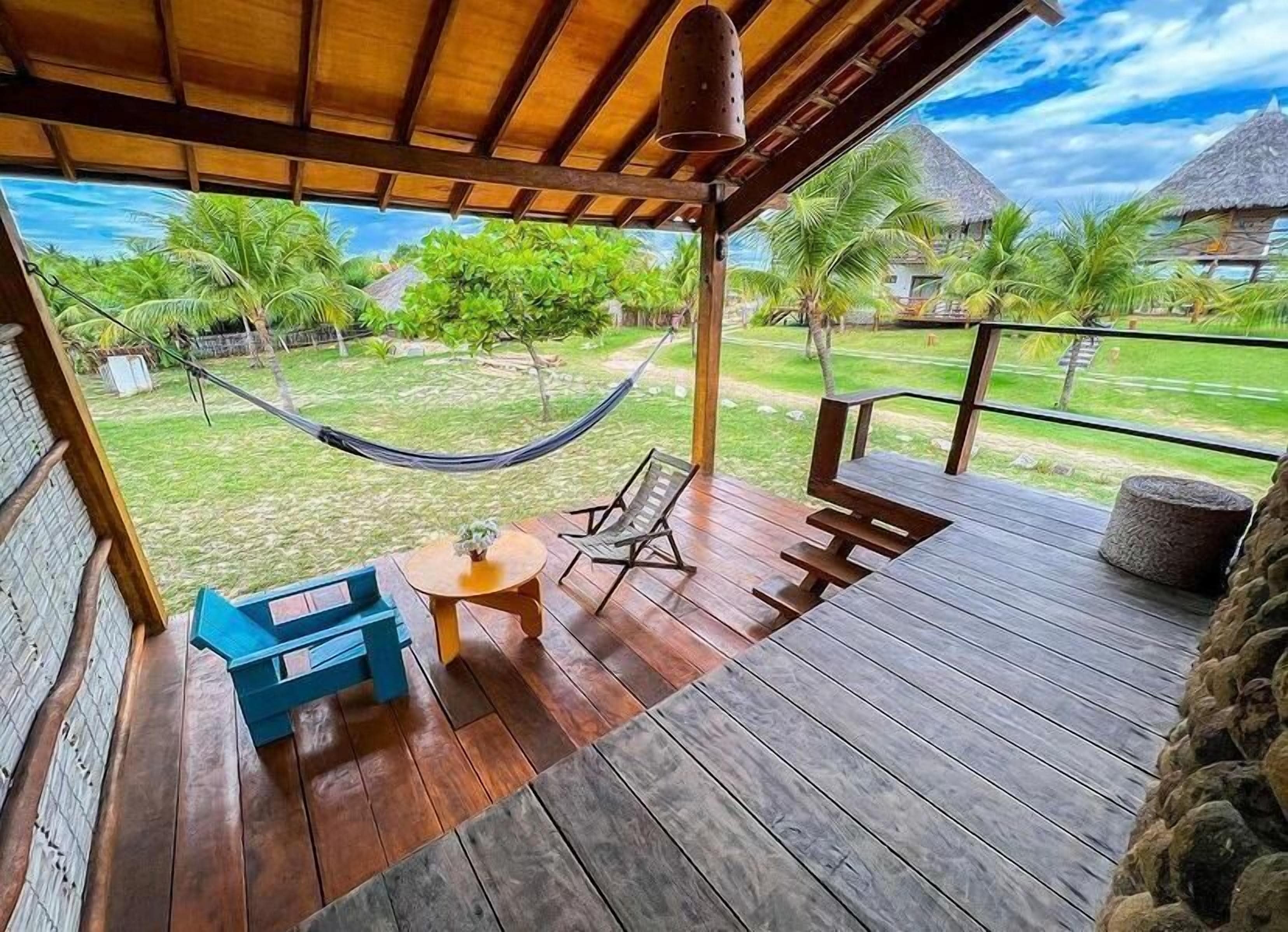 A wooden deck with a hammock, a table, and chairs, overlooking a lush garden with palm trees and a thatched-roof structure.