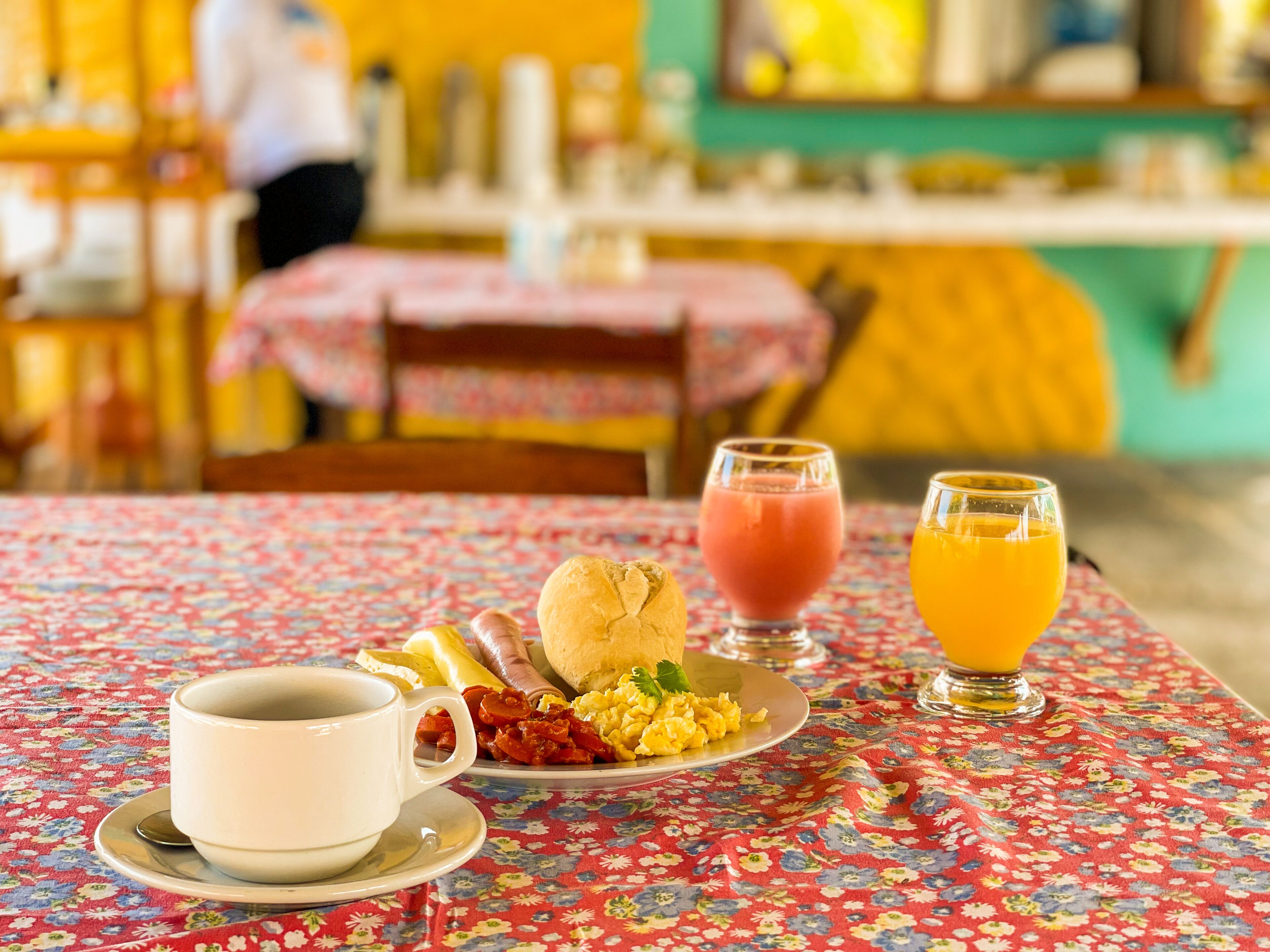 Un desayuno con mantel floreado, una taza, un plato con huevos, tocino y una salchicha, además de dos vasos de zumo de naranja.