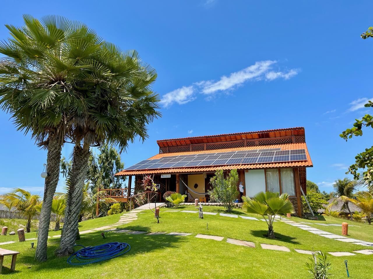 Un edificio con techo de tejas rojas y paneles solares, rodeado de un jardín bien cuidado con palmeras y un cielo azul despejado.