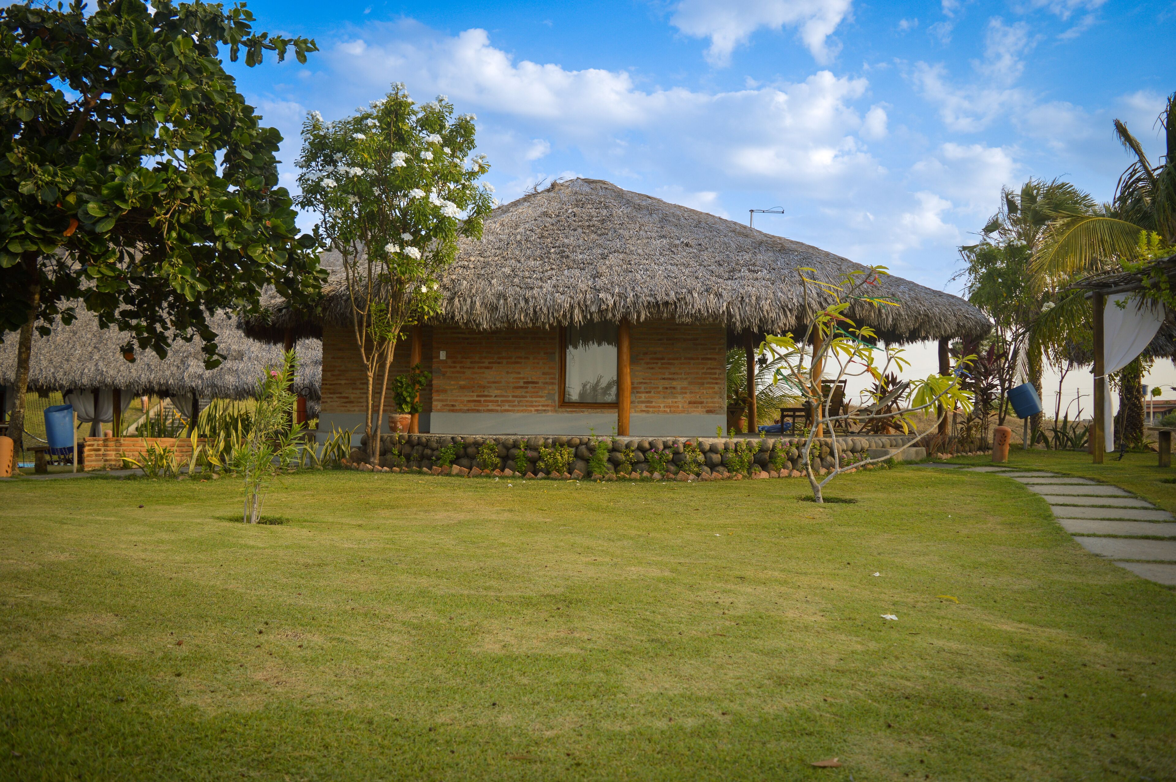 Una casa con techo de paja y fachada de ladrillo, rodeada de un césped cuidado y plantas tropicales.