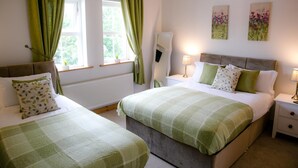 Et soveværelse med to senge, grønne gardiner, et natbord med lampe og blomstermotiver på væggen.