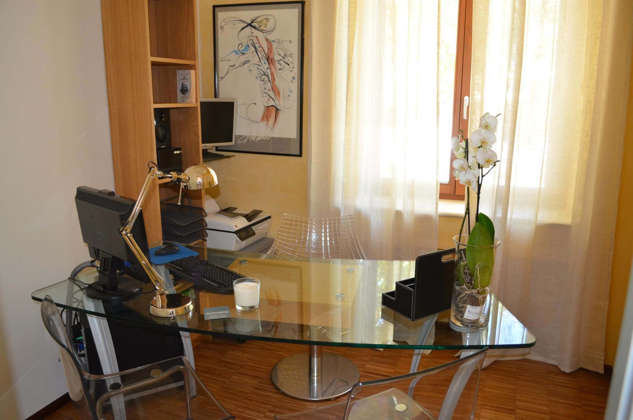 A modern office with a glass desk, computer, and a vase with orchids.