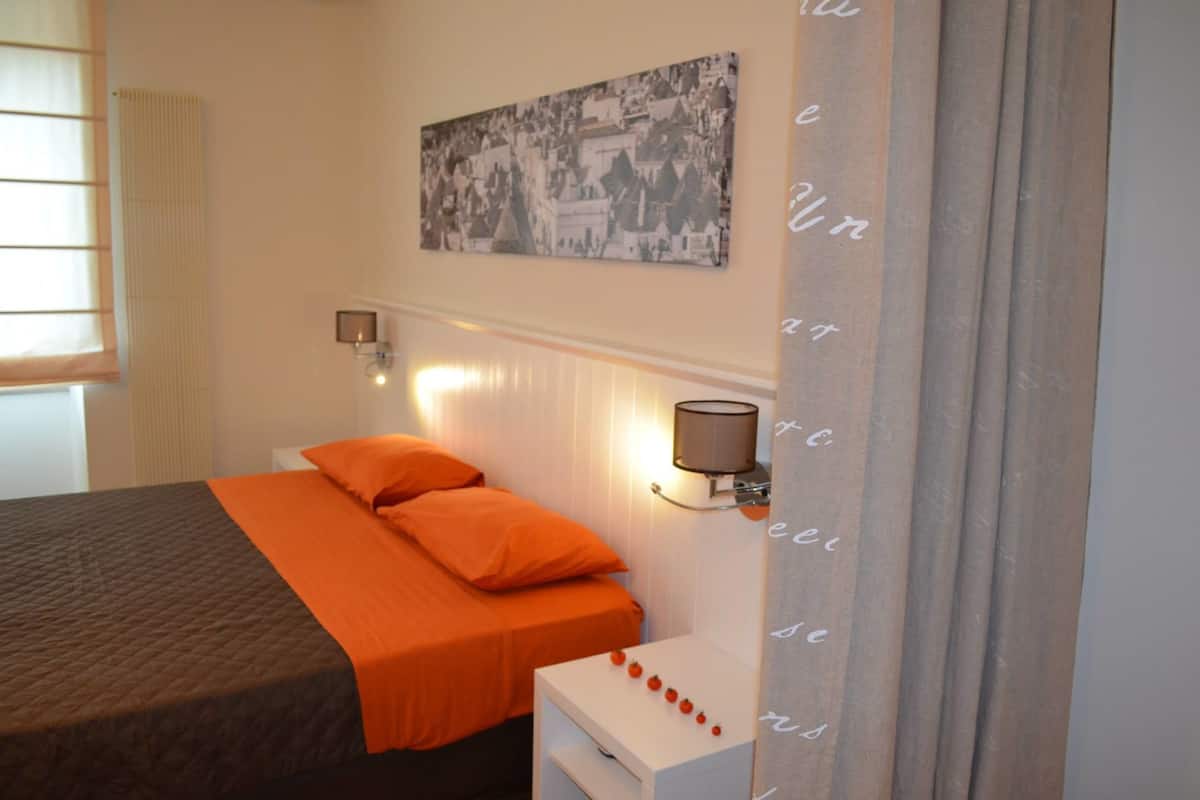 Un dormitorio con cama, ropa de cama naranja, una mesita de noche, una lámpara de pared y un cuadro enmarcado sobre la cama.