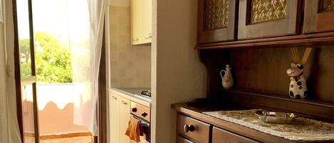 Una cocina con armarios de madera, suelo de baldosas y vistas a un balcón por una puerta abierta.