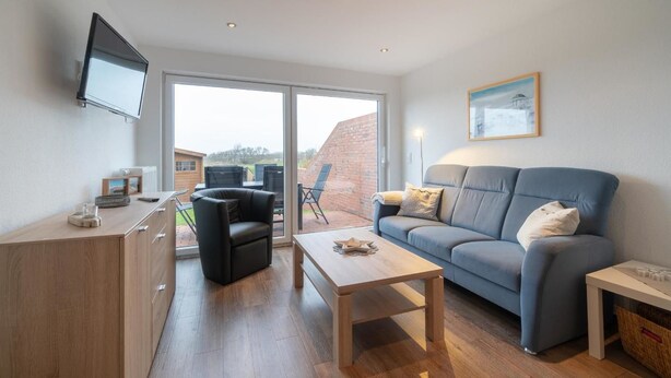 A modern living room with a sofa, a coffee table, a TV mounted on the wall, and a large sliding glass door leading to an outdoor patio. - Reede Hus Alfonso (Borkum)