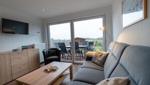 A modern living room with a sofa, a coffee table, a TV mounted on the wall, and a sliding glass door leading to an outdoor patio area. - Reede Hus Alfonso (Borkum)