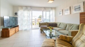 En moderne stue med et fladskærms-tv, tv-møbel i træ, glasbord og en stribet sofa, samt udsigt ud gennem en glasdør.