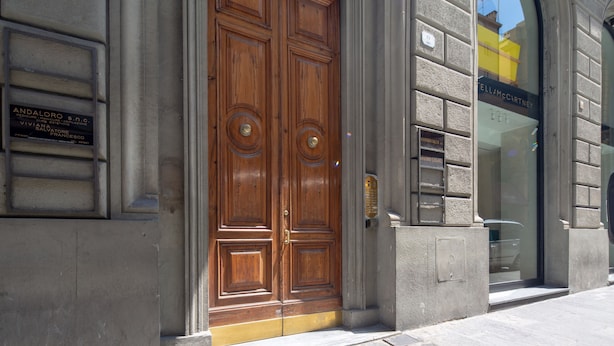 A double wooden door with a plaque, a stone wall, and a clear sky visible through a window. - Elegant and comfortable apartment near the Duomo. The perfect location. (Firenze)