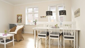A dining area with a white table and chairs, a wicker chair, a TV, and artwork on the walls. - Aalborg City apartments (Aalborg)