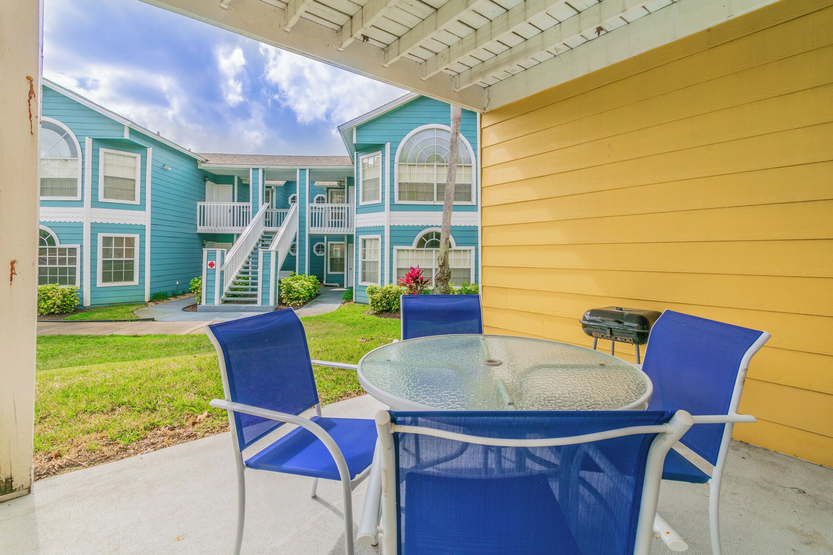 A covered patio with a glass table and blue chairs, a yellow wall, and a blue building with a staircase in the background.