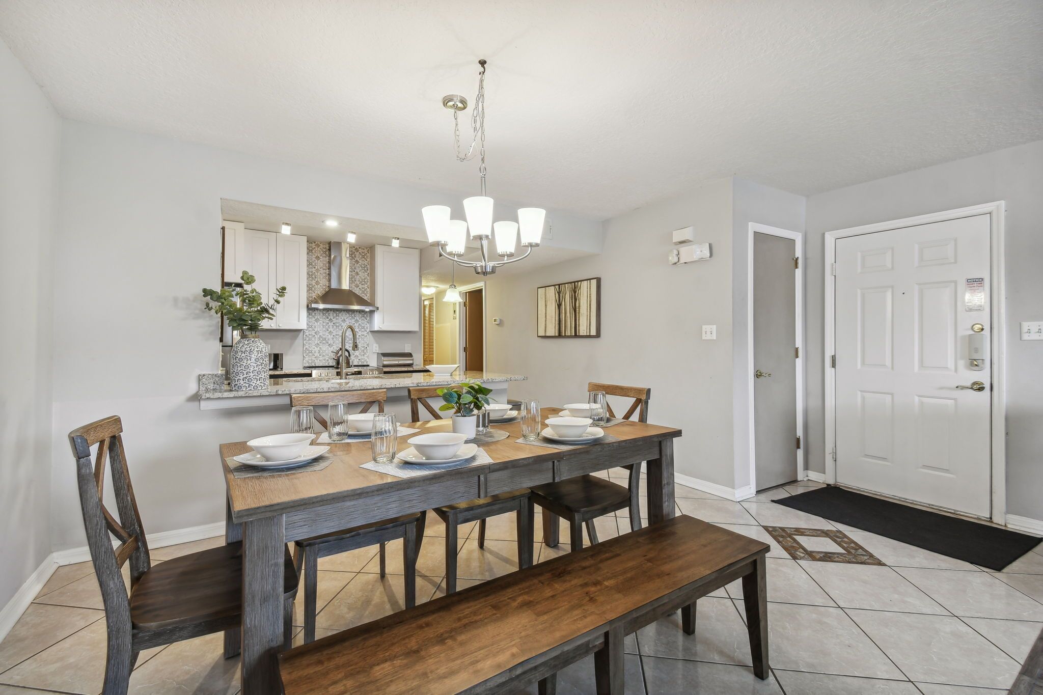A dining area with a wooden table set for four, a chandelier, and a kitchen in the background.
