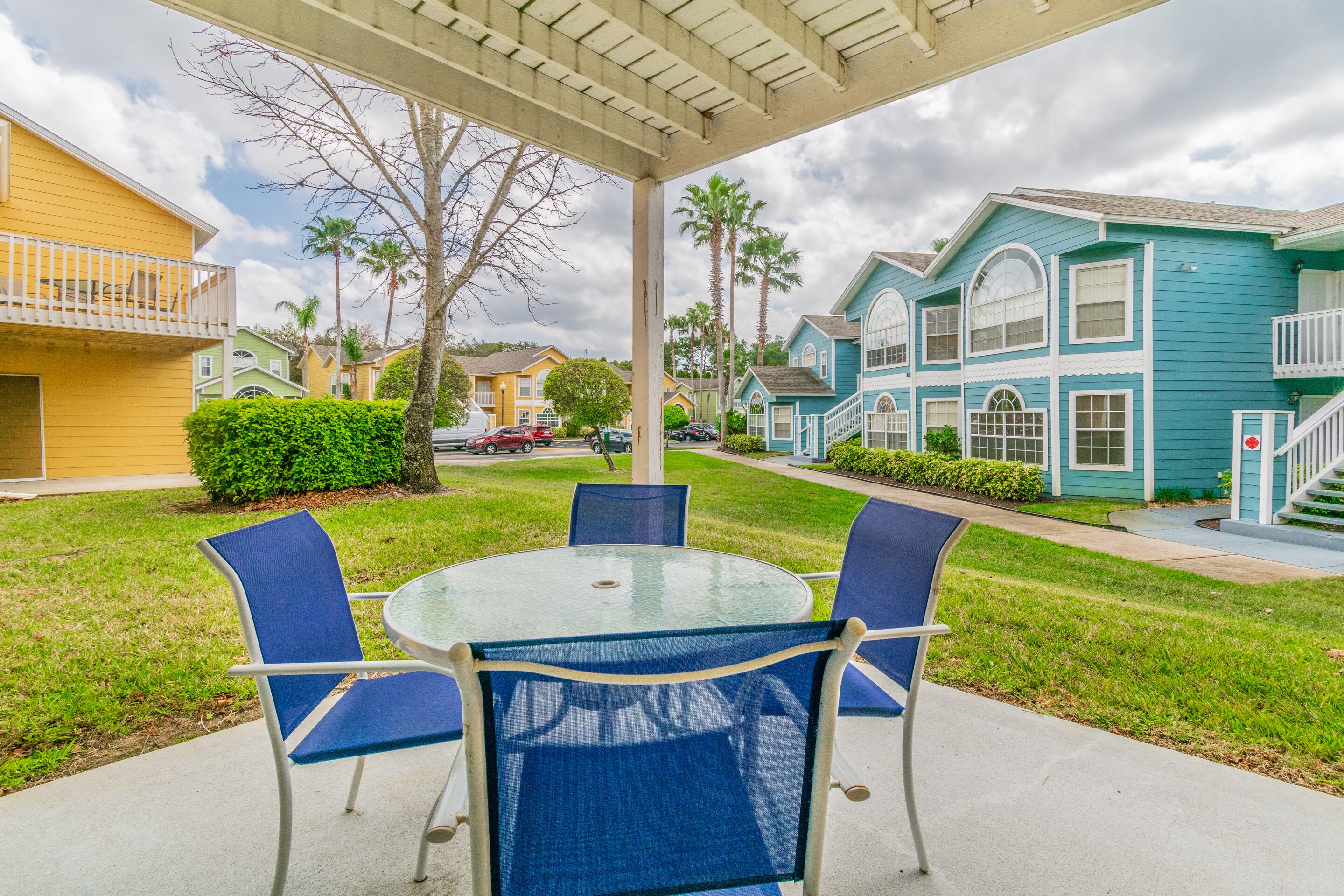 A covered patio with a round glass table and blue chairs, surrounded by well-maintained lawns and colorful houses.