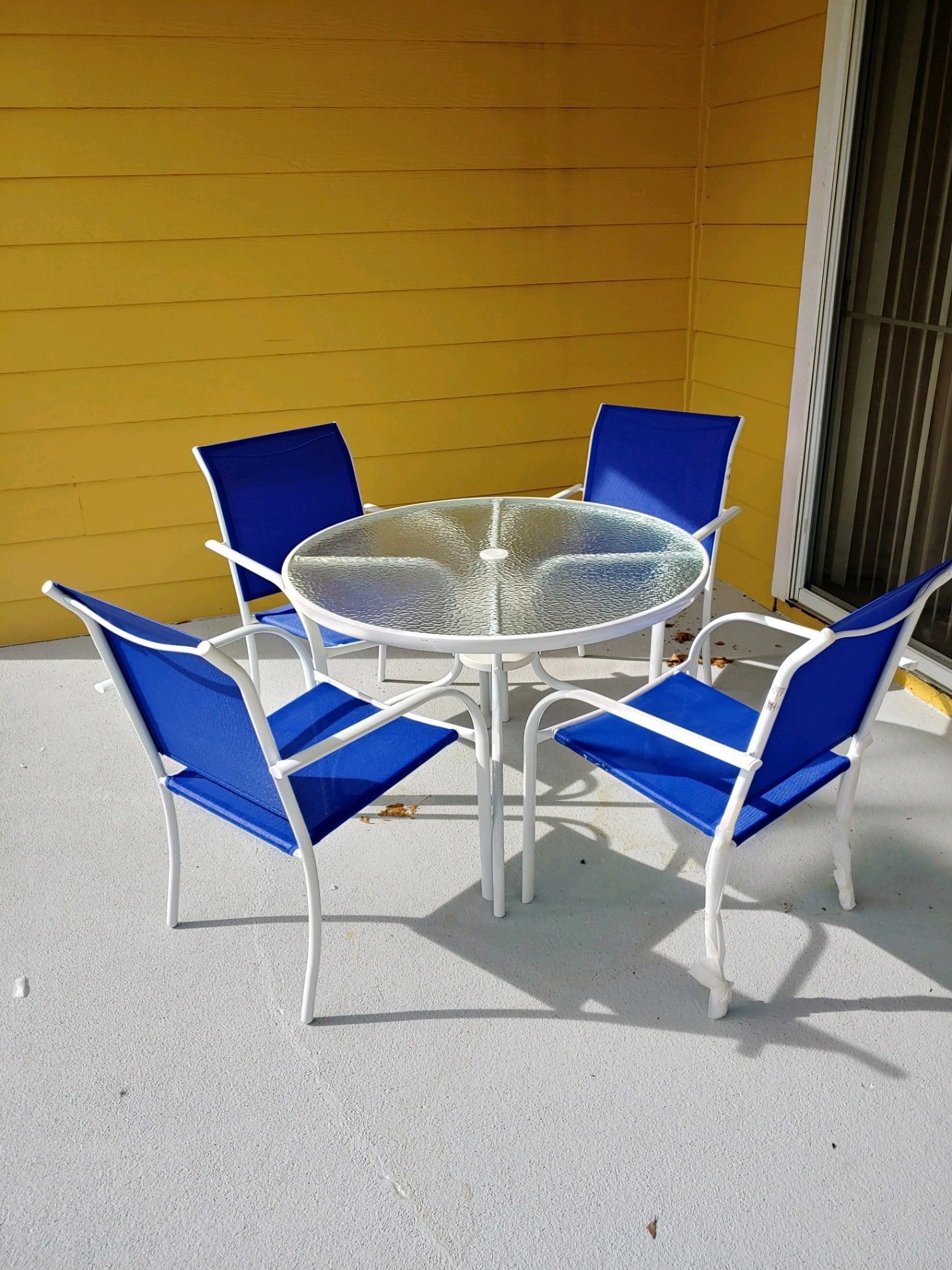 A round glass table with four blue chairs, set against a yellow wall.