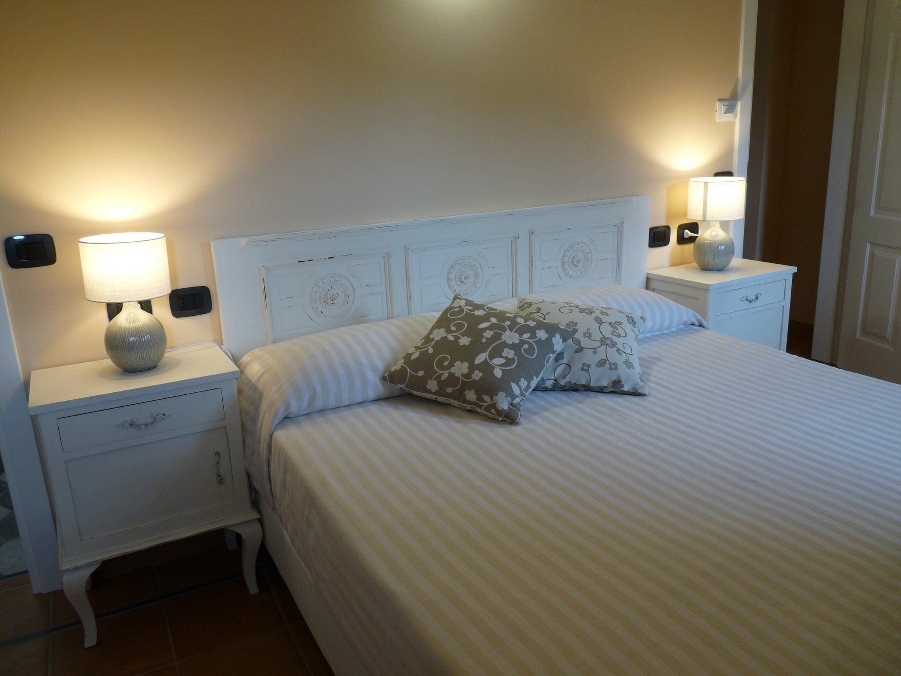 A neatly made bed with striped bedding, two bedside lamps, and a white nightstand with a drawer.