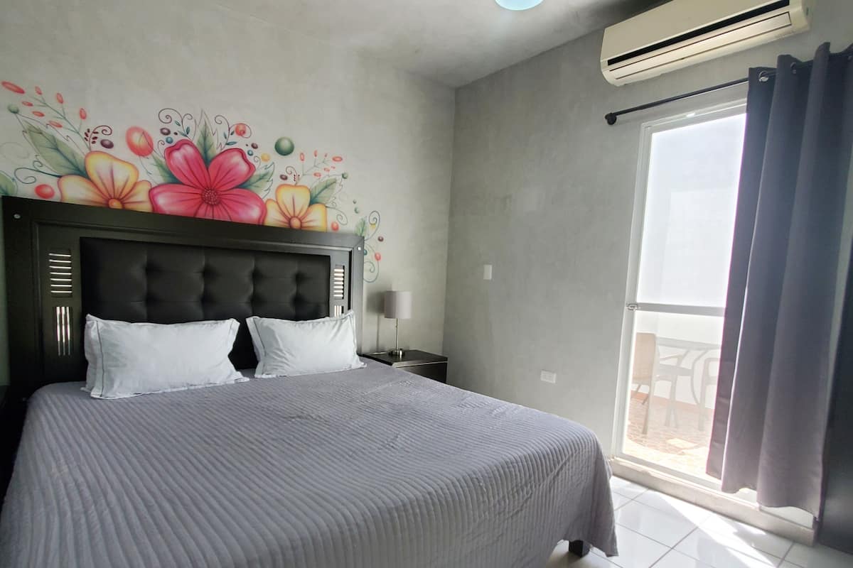 Ein Schlafzimmer mit einem großen Bett, einem Kopfteil mit vertikalen Lamellen, einem Nachttisch, einer Wandleuchte und einem Fenster mit Vorhängen.